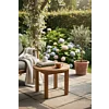 Deze massief houten bijzettafel vormt het middelpunt van een serene buitenruimte, ideaal voor een moment van rust met koffie en een boek. De warme houttinten harmoniëren prachtig met weelderige tuinbeplanting, wat een luxe en natuurlijke sfeer creëert.